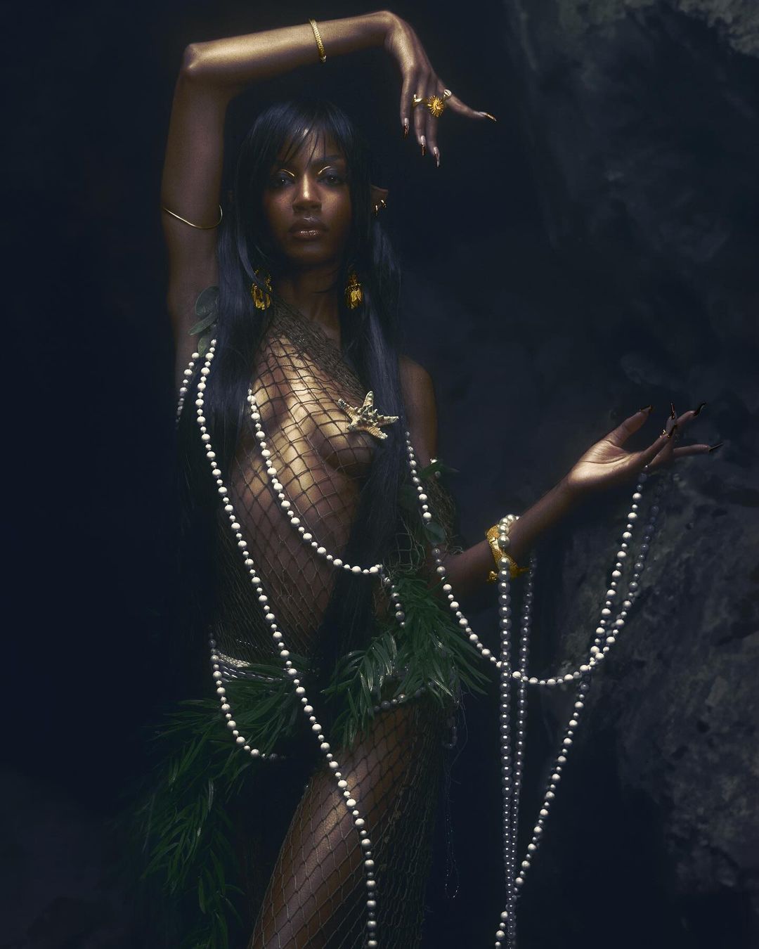 Glamorous dark-skinned woman with long hair posing in a fishnet garment adorned with greenery and pearls in a dark, cave-like setting.