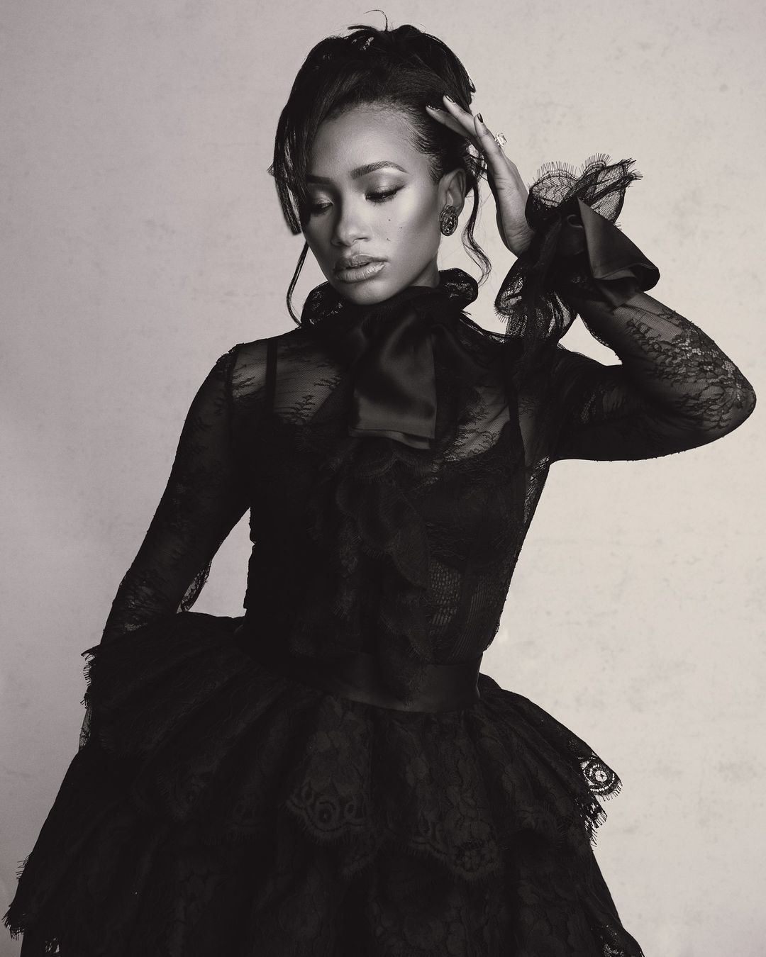 Monochrome portrait of a woman wearing a black lace dress with a high neckline and ruffled sleeves, posing in front of a neutral backdrop.