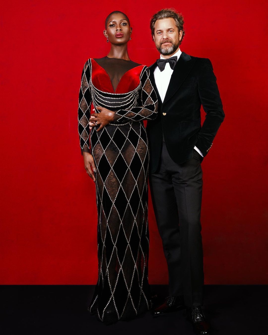 Elegant portrait of a Black woman in a long gown and a Caucasian man in a tuxedo, posing against a vibrant red backdrop in a studio.