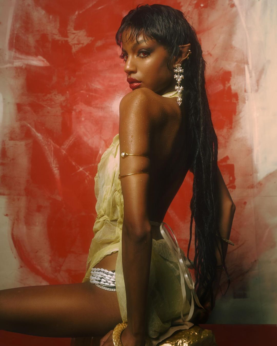 A dark-skinned woman with pointed ears, back turned to the camera, poses in a yellow dress against a red and white abstract backdrop.