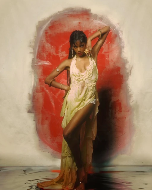 Elegant Black woman poses in yellow dress with ornate bracelets and earrings in a studio setting with red and white painted backdrop.