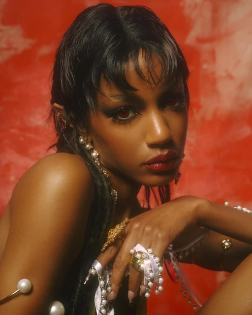 Portrait of a woman with dark hair and dramatic red makeup wearing pearl jewelry against a red-and-white textured background.