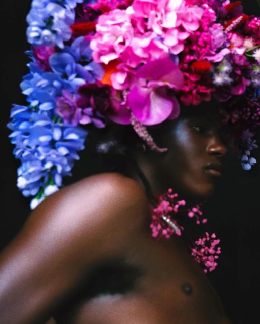 Artistic close-up of a Black woman with a colorful floral headpiece and a flower necklace, set against a dark backdrop.