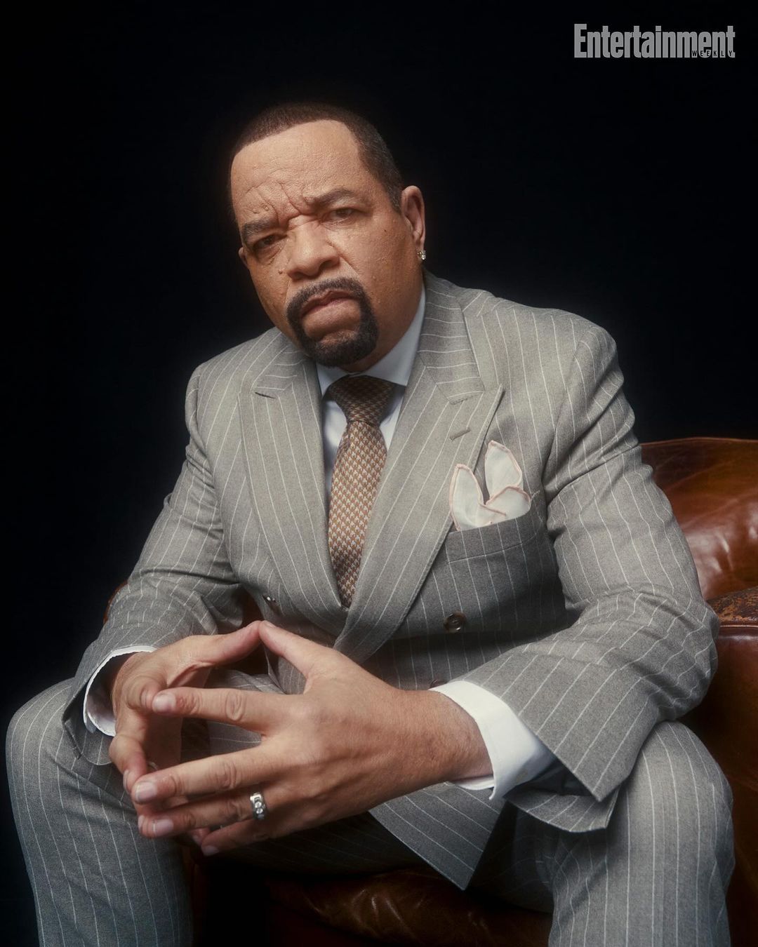 Portrait of Ice-T wearing a pinstripe suit and tie, seated against a dark background, for Entertainment magazine editorial.