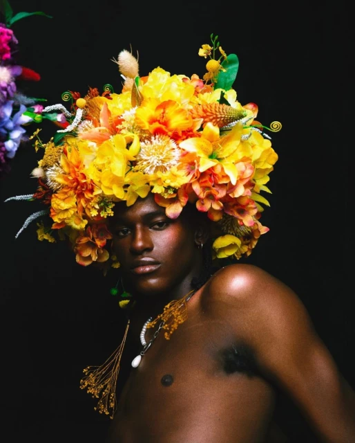 Portrait of a shirtless Black man wearing a vibrant floral headpiece, set against a dark backdrop.