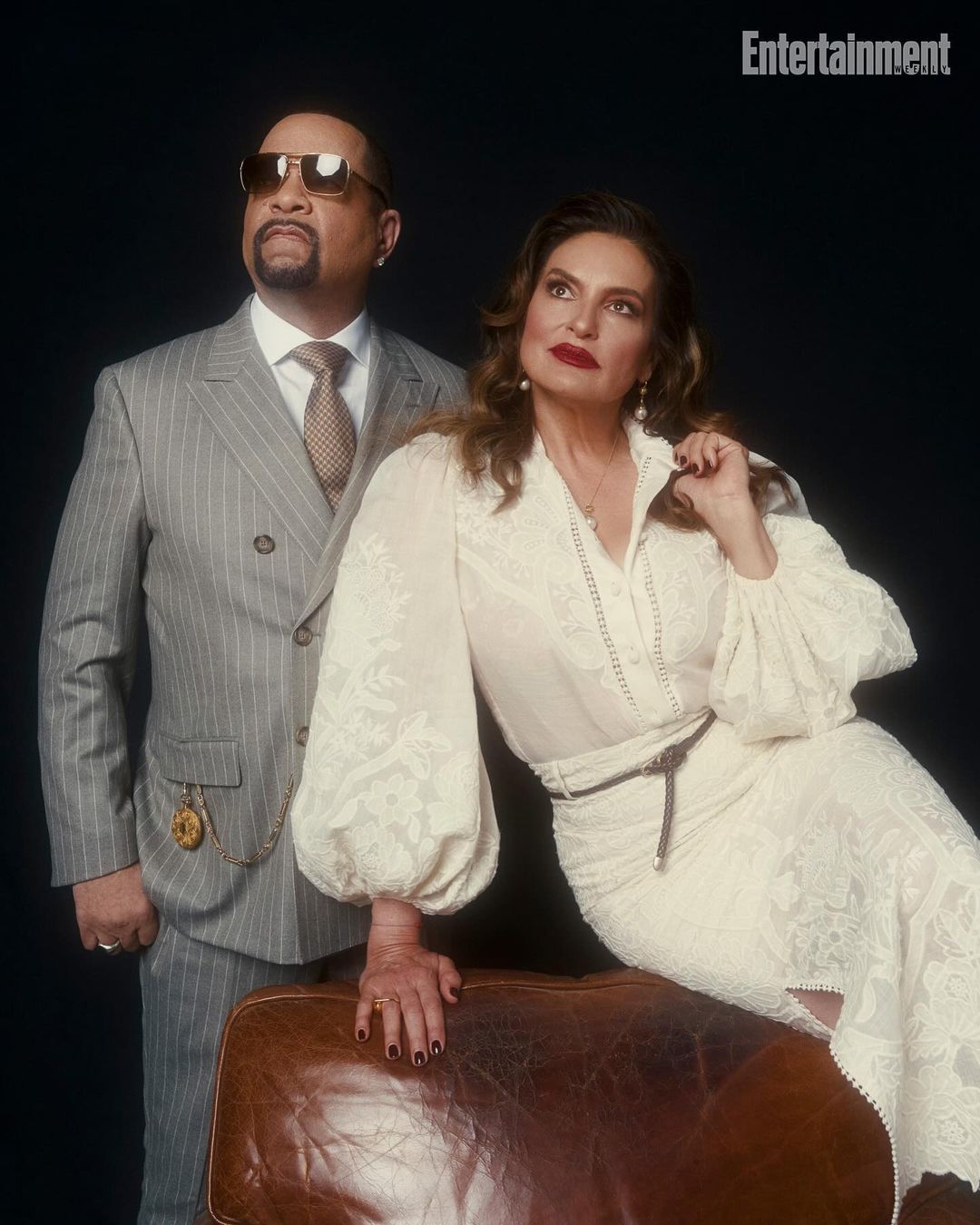 Portrait of Ice-T in a grey suit and Coco Austin in a white lace dress, posed against a dark backdrop with a leather trunk.