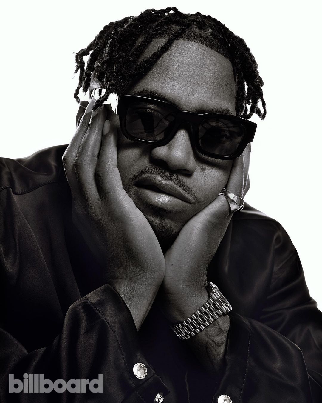 Monochrome portrait of a stylish man with dreadlocks, sunglasses, a ring, and a watch, holding his face against a bright white backdrop.