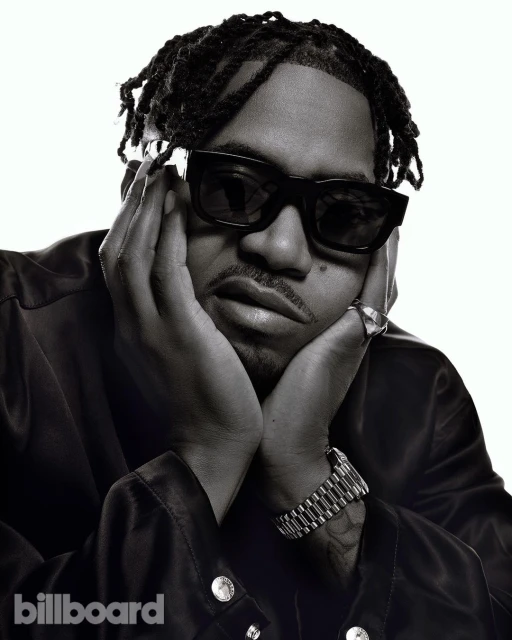 Monochrome portrait of a stylish man with dreadlocks, sunglasses, a ring, and a watch, holding his face against a bright white backdrop.