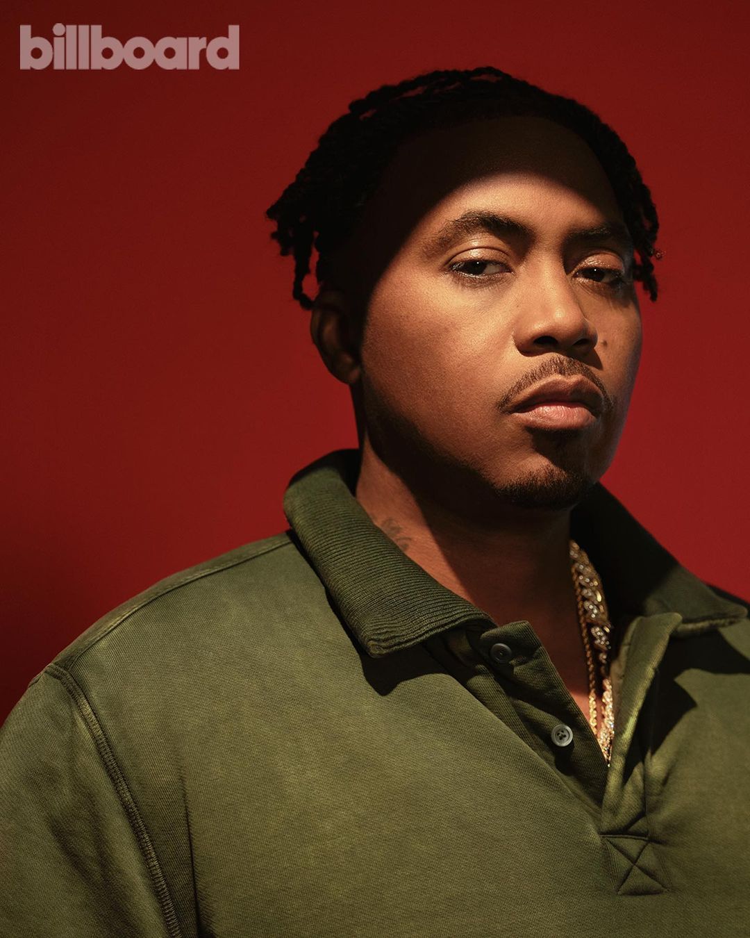 Portrait of the American rapper Nas in an olive green polo shirt with a gold chain necklace against a solid red background.