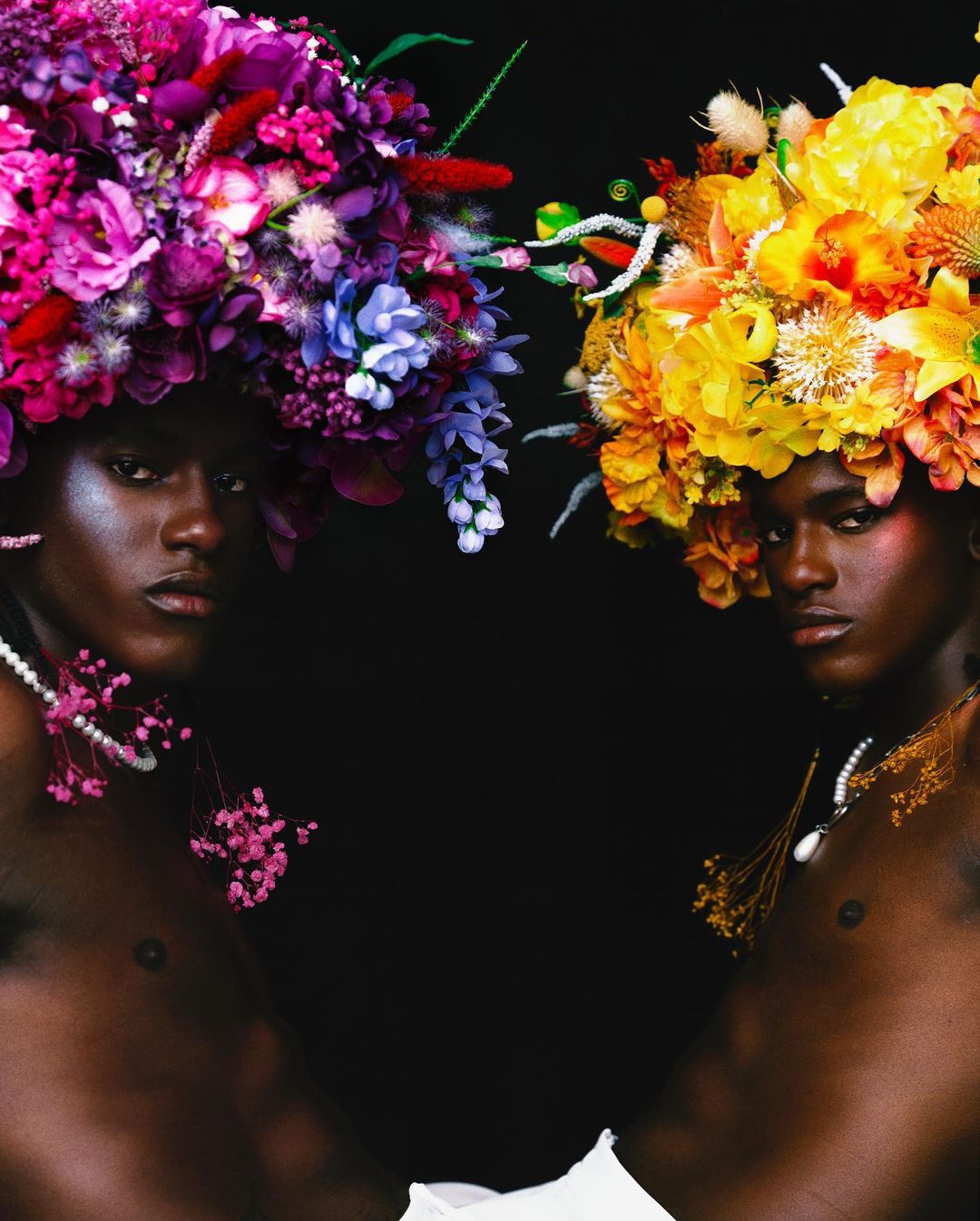 Two models with dark skin pose in a studio, wearing vibrant floral headpieces and pearl necklaces against a stark black backdrop.