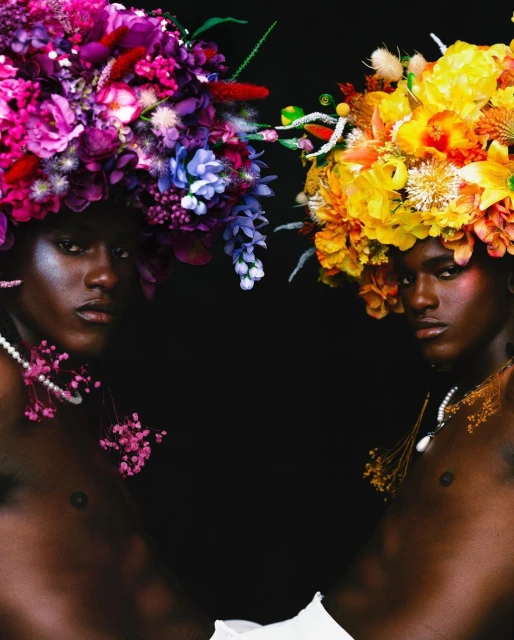 Two models with dark skin pose in a studio, wearing vibrant floral headpieces and pearl necklaces against a stark black backdrop.