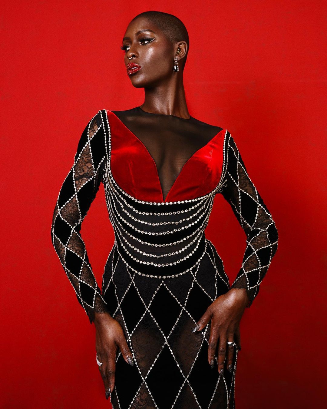 Elegant Black woman in a black velvet gown with diamond accents poses against a solid red backdrop in a fashion studio shot.
