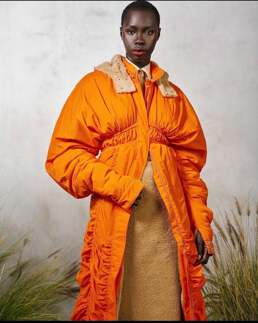 A dark-skinned fashion model in an orange puffer coat is posing in front of a textured wall with plants at the bottom of the image.