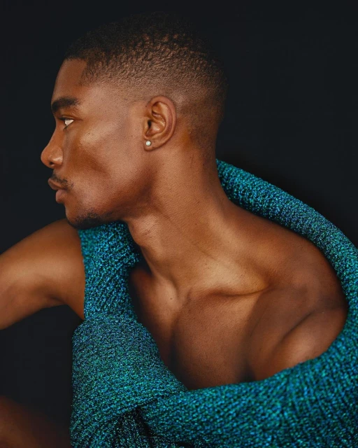 Close-up studio shot of an African American male model with short hair wearing a turquoise knit top against a dark background.