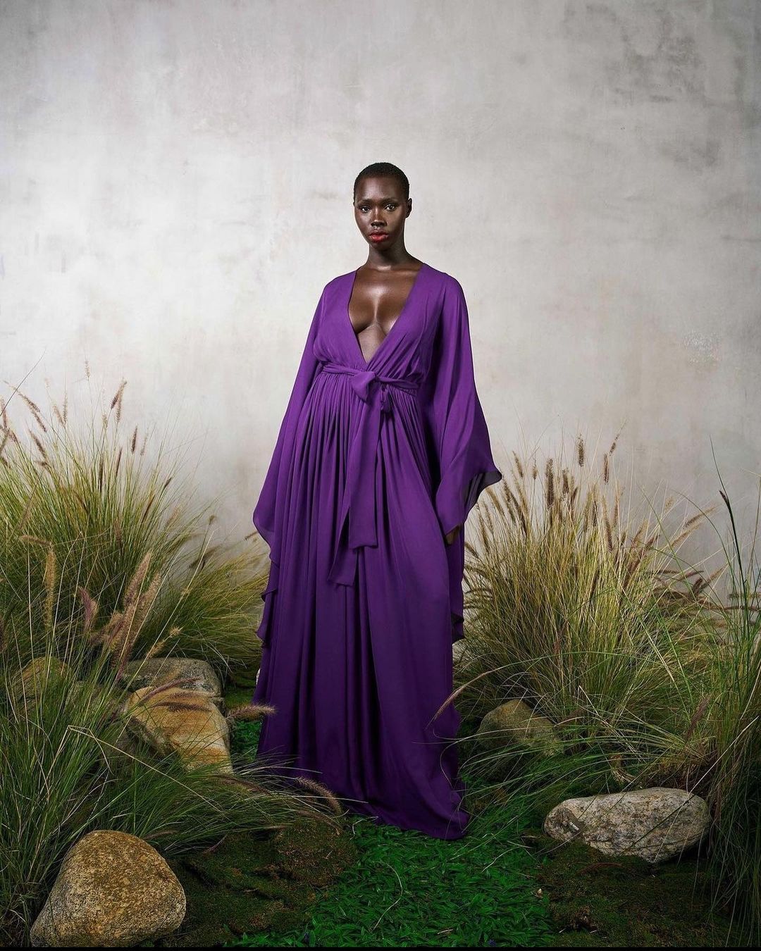 Woman in elegant purple dress posing in natural setting with tall grasses and textured gray background for fashion editorial shoot.