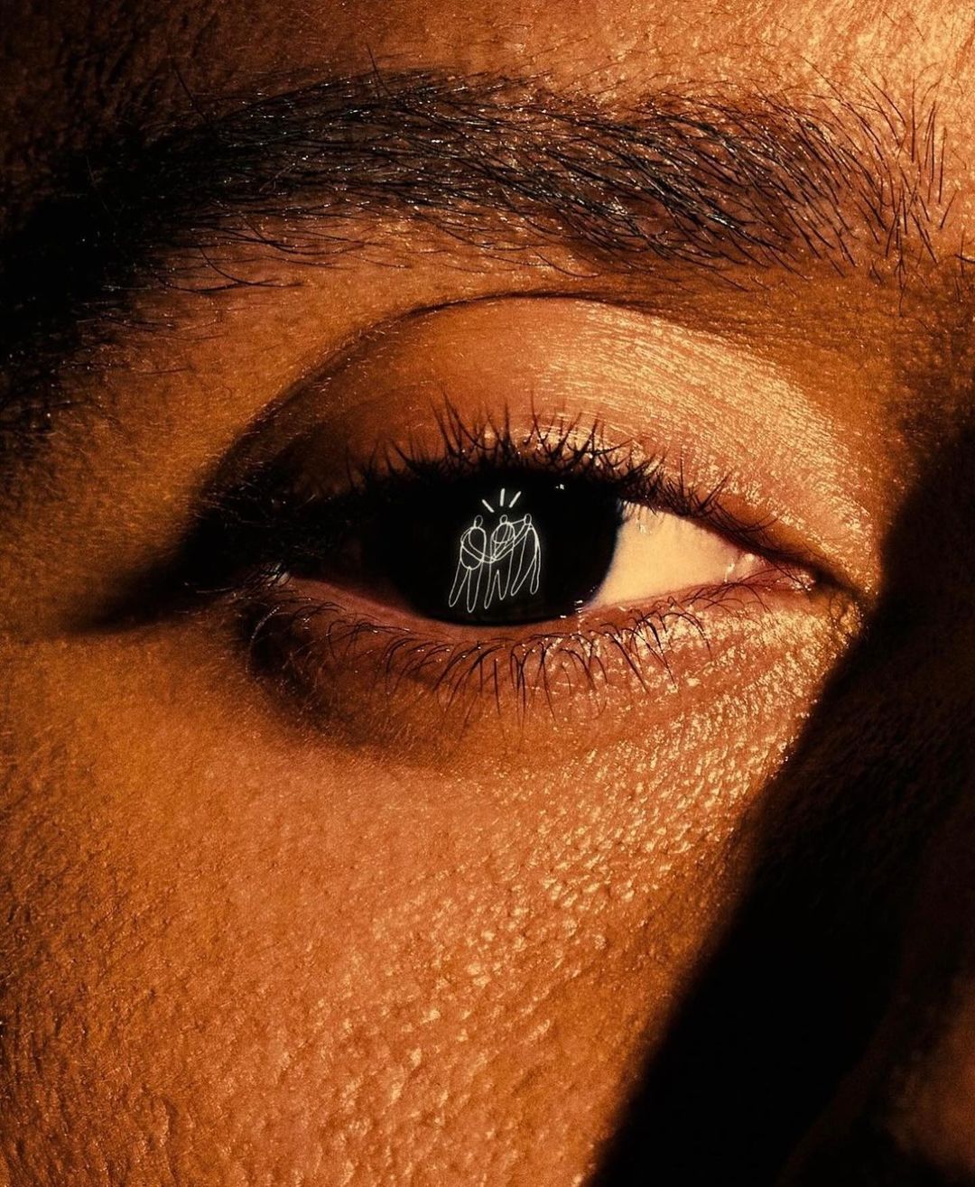 Macro shot of a dark-skinned person's eye reflecting a small illustration of three figures, capturing detail and texture in warm lighting.