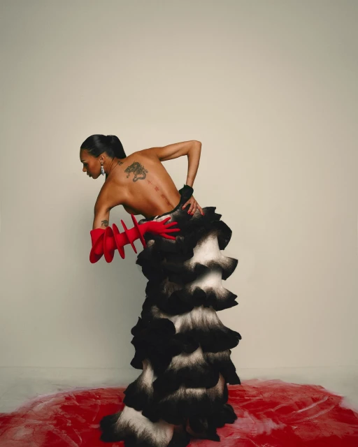 Fashion portrait of a topless adult woman in a black and white ruffled dress with red glove-like sleeves, posing against a white backdrop.