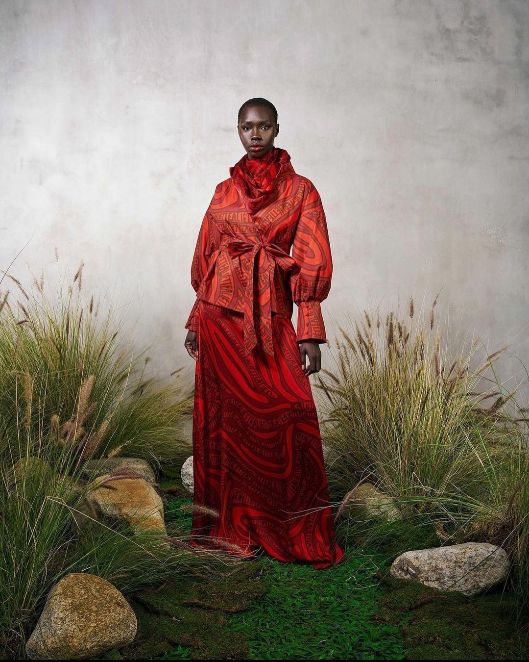 A woman in a patterned red dress and jacket stands in an outdoor setting with tall grasses and rocks, creating a fashion editorial image.