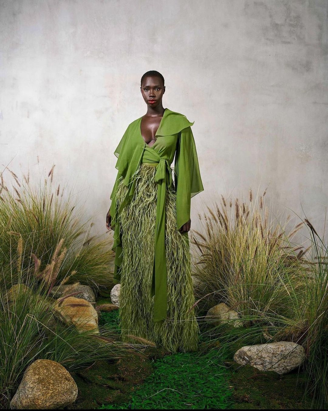 An African-descent woman in an olive-green gown with feathered details poses in a studio with a background of tall grass and mossy rocks.