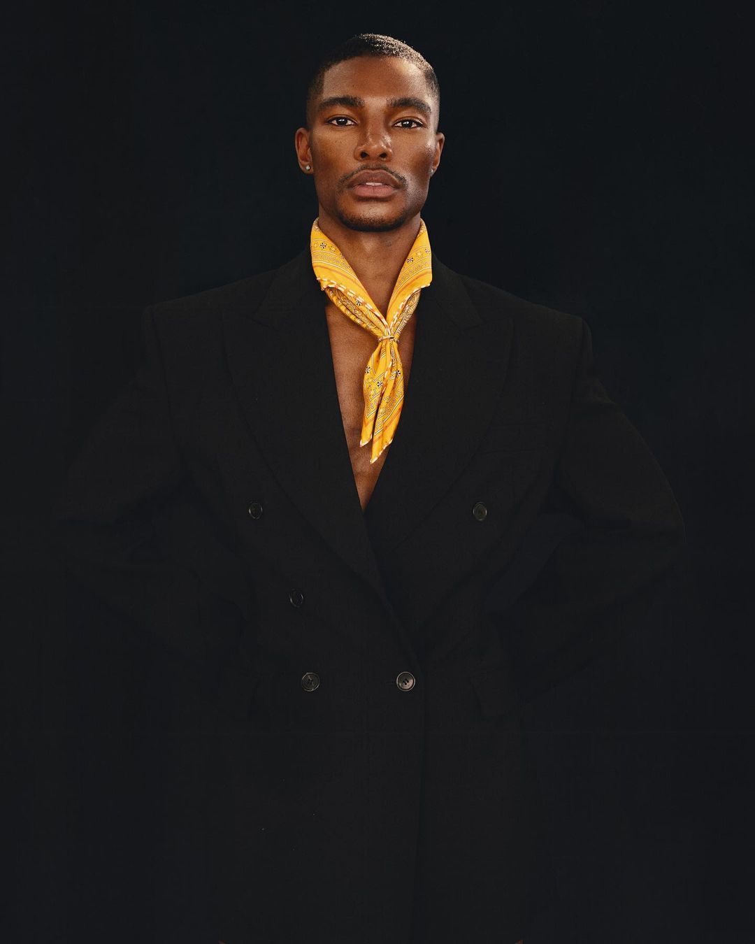 Portrait of a Black male model wearing a double-breasted black jacket and a yellow paisley scarf around his neck, against a dark backdrop.