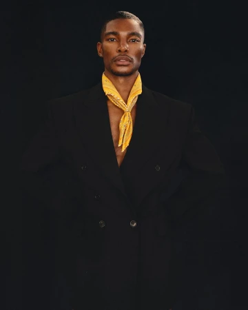 Portrait of a Black male model wearing a double-breasted black jacket and a yellow paisley scarf around his neck, against a dark backdrop.
