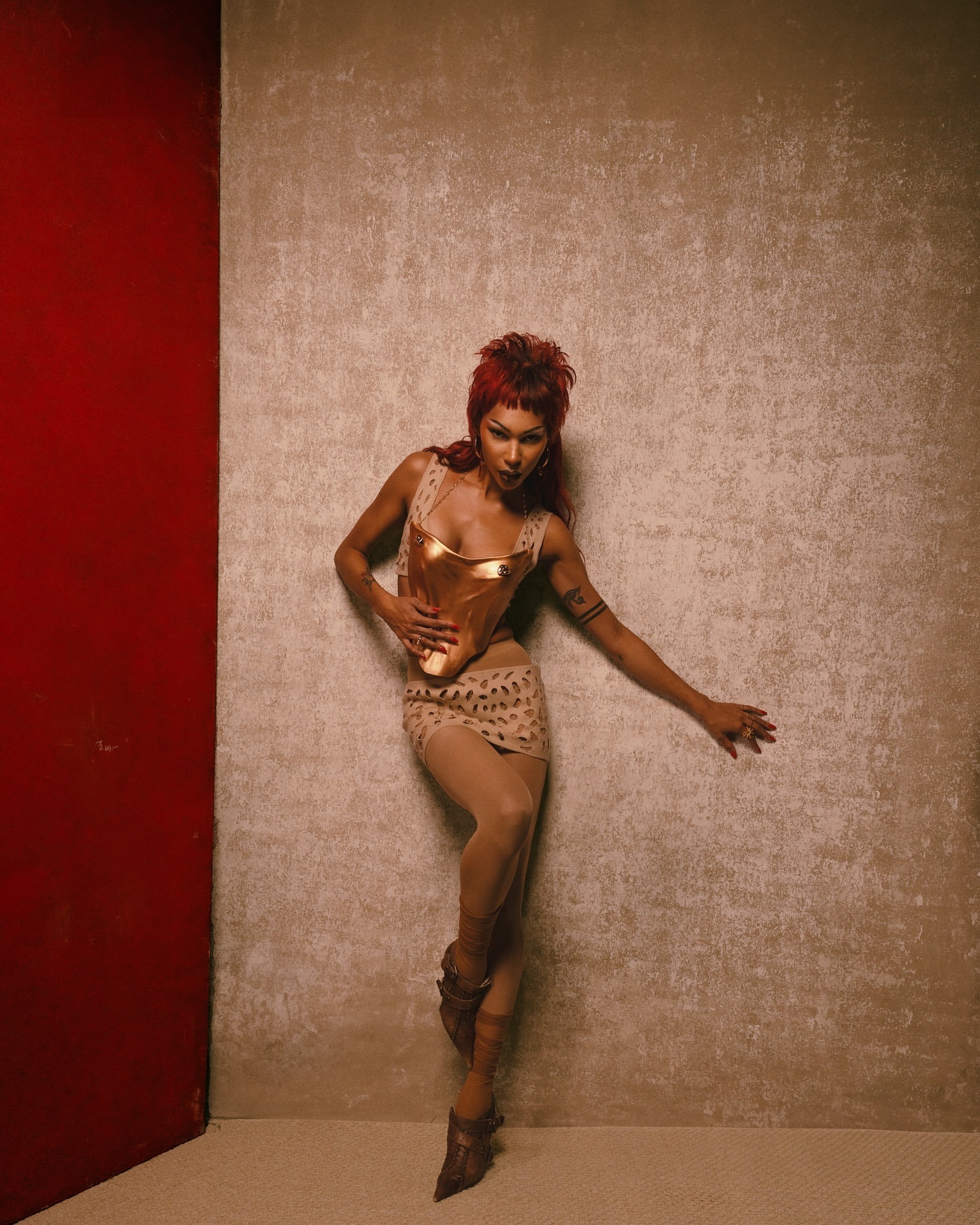 A woman with red hair poses in a gold top and skirt against a textured backdrop, highlighted by a vibrant red wall section.