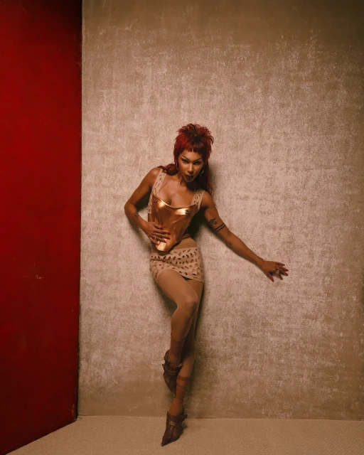A woman with red hair poses in a gold top and skirt against a textured backdrop, highlighted by a vibrant red wall section.