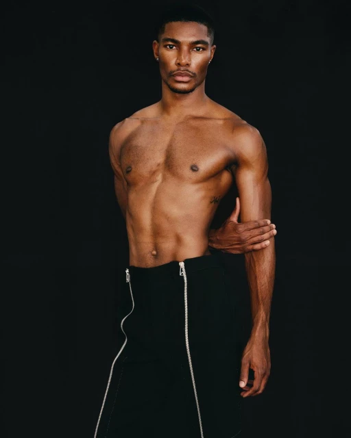 Striking studio portrait of a muscular, shirtless African American male model in black pants with silver zippers against a black background.
