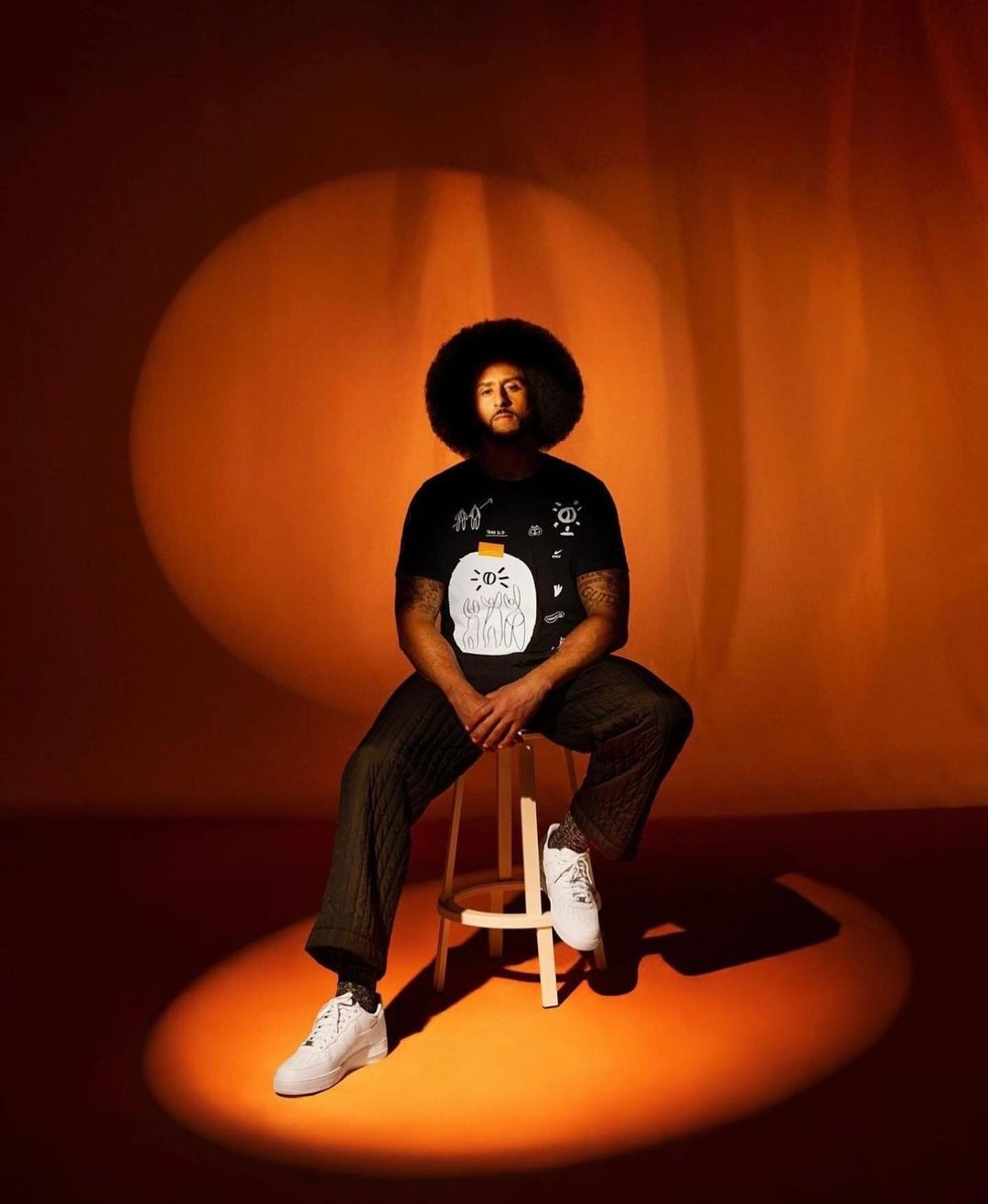 A man with an afro wearing a graphic tee sits on a stool in a studio. He is illuminated by two spotlights on an orange background.