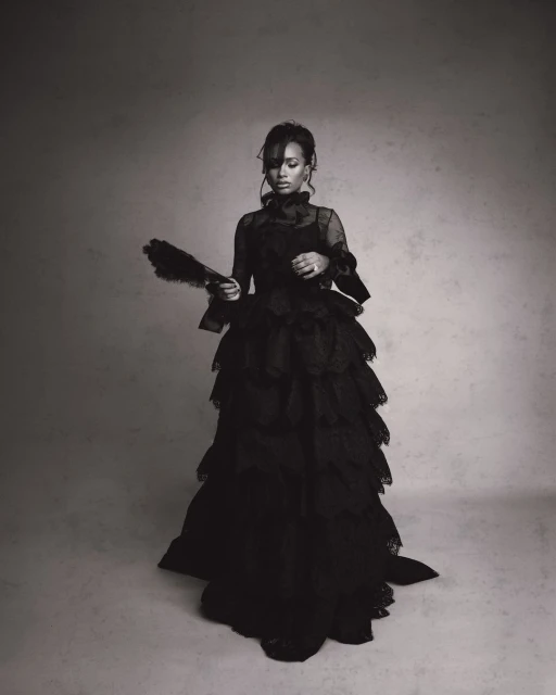 An adult female poses in a dramatic, layered black gown while holding a fan, captured in a black and white studio portrait.
