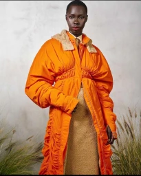 Black woman wearing a bright orange puffer coat with tan shearling collar and textured tan skirt