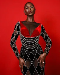 Black woman with a shaved head wearing a black and red harlequin patterned dress against a red backdrop