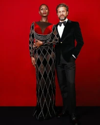 Portrait of a bald black woman in a long black and red patterned gown standing beside a man in a black velvet tuxedo by Obidigbo Nzeribe