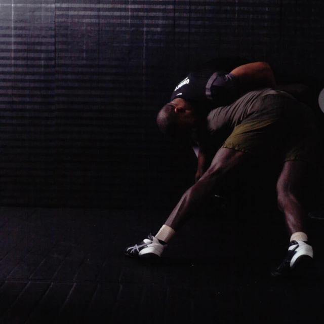 Two adult males engaged in a wrestling match, grappling indoors on a dark floor with a horizontally textured wall in the background.