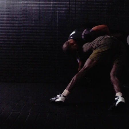 Two adult males engaged in a wrestling match, grappling indoors on a dark floor with a horizontally textured wall in the background.