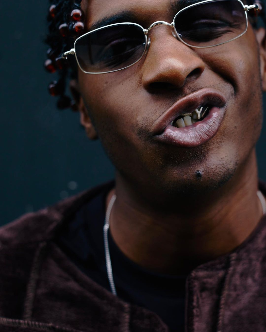 Close-up portrait of an adult with dark hair, wearing brown suede jacket, silver chain necklace, and rimless rectangular sunglasses. 