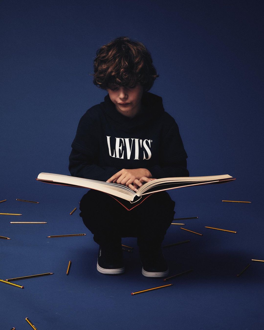 Young boy reading an open book in a photo studio with pencils on the floor and a blue background, wearing a Levi's hooded sweatshirt.
