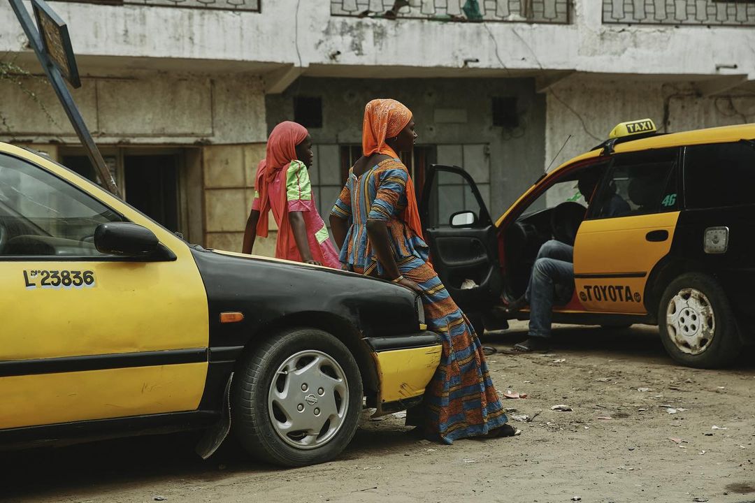 Two women in colourful dresses sit on a yellow and black cab in a city street, with a second cab visible in the background.