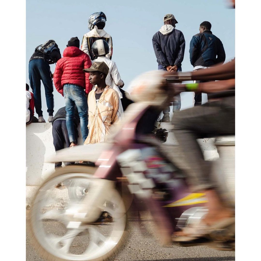 Outdoor shot of a group of people, including one riding a motorbike with motion blur, on a sunny day near a concrete barrier.
