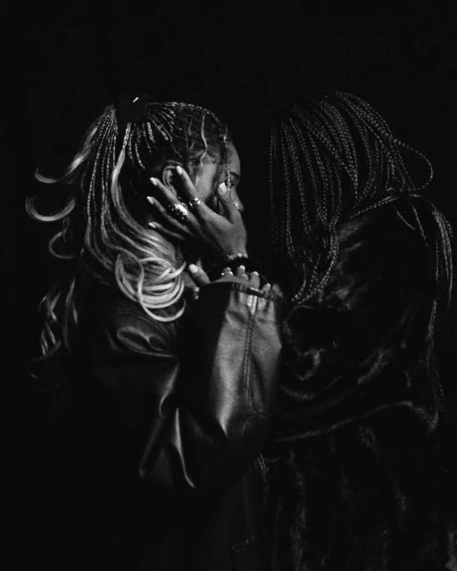 Black and white portrait of two African American women with braided hair in leather jackets, face to face in a dimly lit, close-up shot.