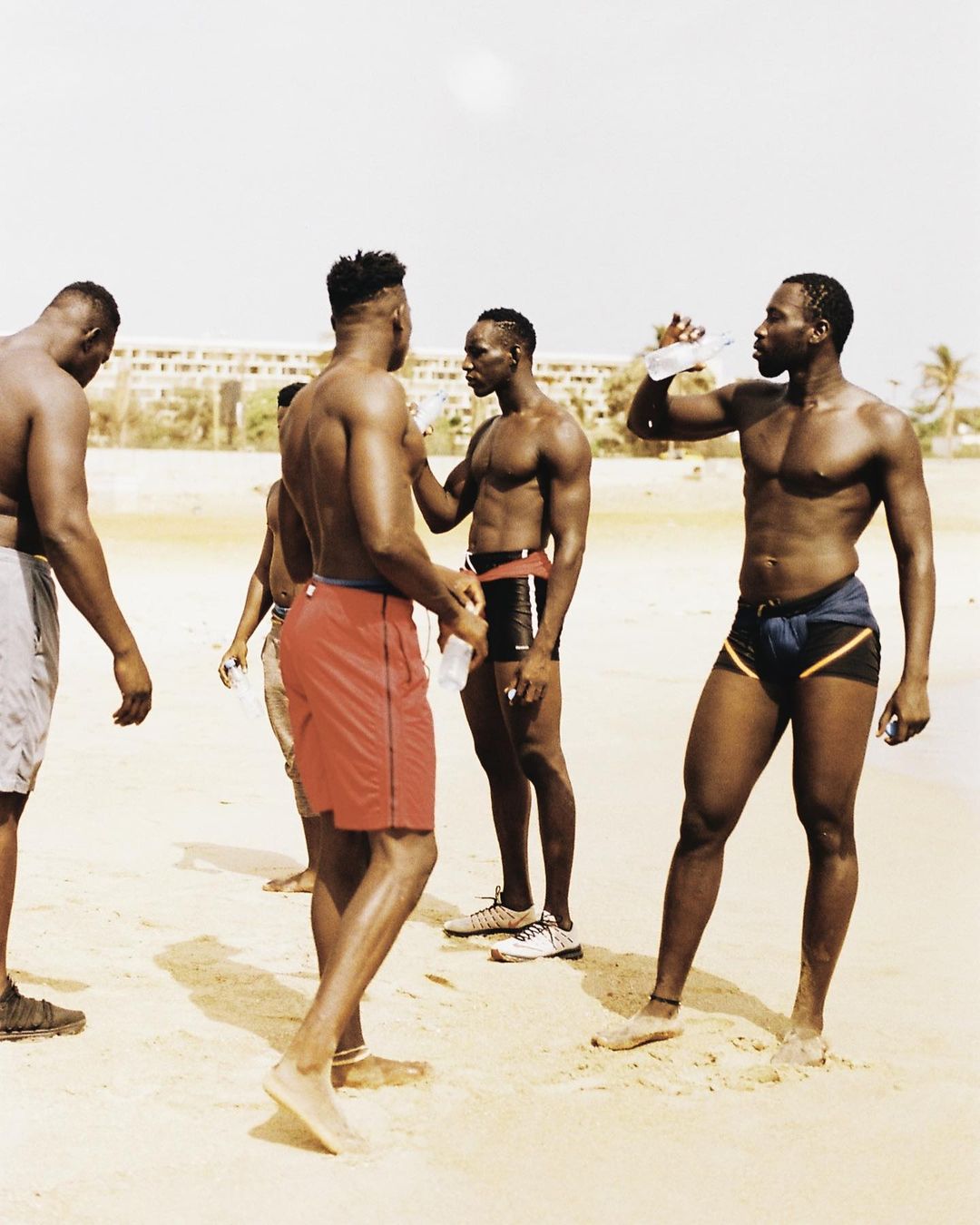 A group of muscular adult men on a sandy beach, some shirtless and drinking water, with a low-rise building visible in the distance.