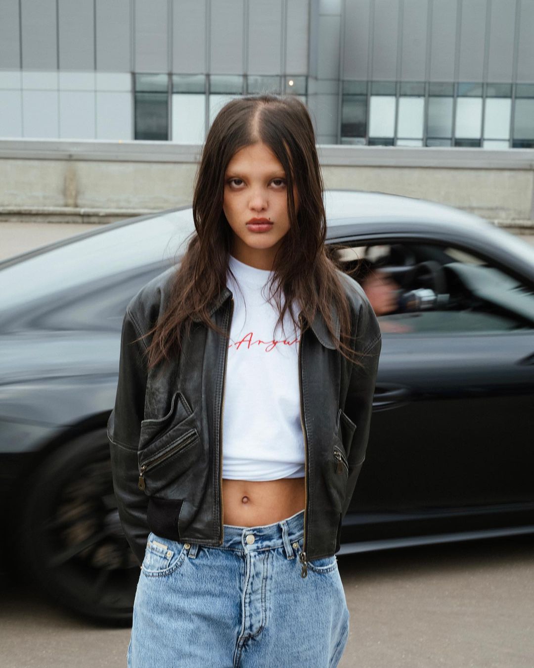 A young woman in a leather jacket and jeans poses in front of a black sports car and a modern building on an overcast day.