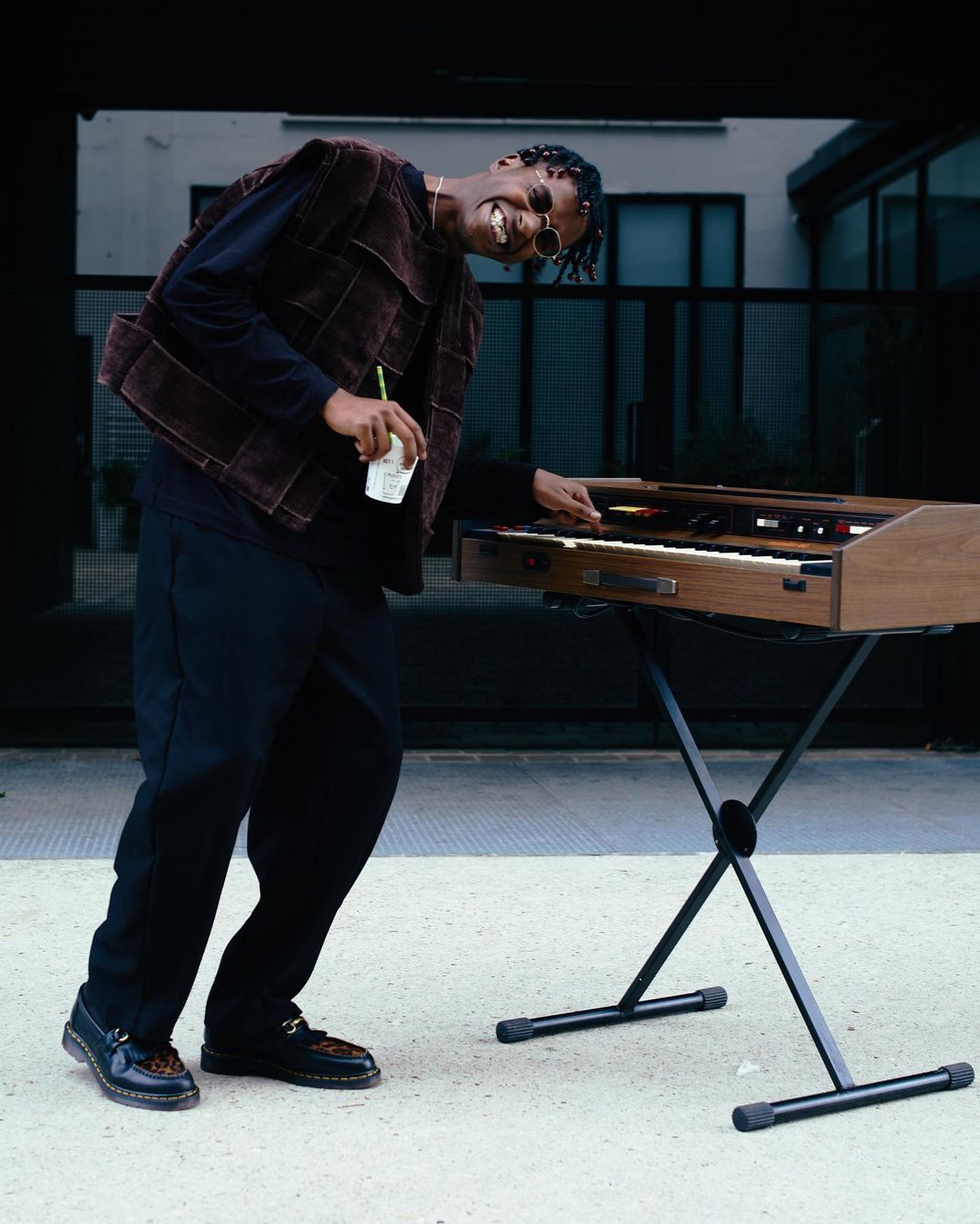 Smiling Black man plays a keyboard outdoors in stylish vest and round sunglasses. Building and grey pavement in the background.