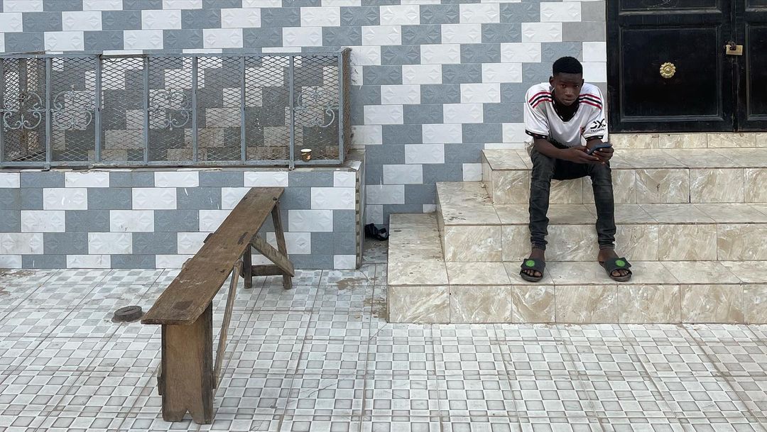 Teenager sits on steps with his smartphone in front of a patterned wall and an old wooden bench in an outdoor urban setting.