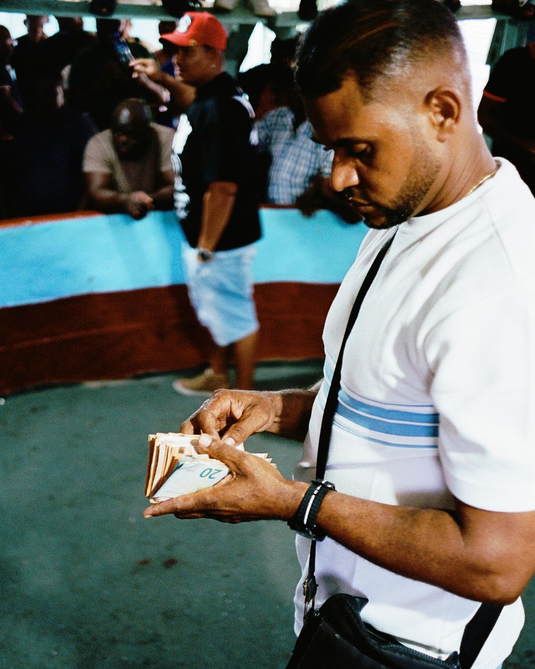 Adult male counts euro notes. The man wears a white shirt with blue stripes and a black shoulder bag. Other people are visible.