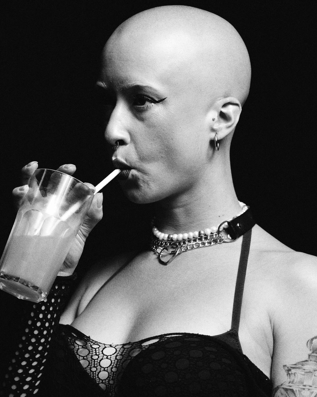 Monochrome portrait of a bald adult female drinking from a glass through a straw, wearing a lace top and pearl choker, with a dark backdrop.
