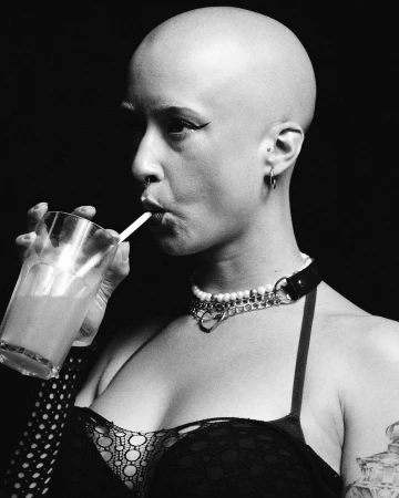 Black and white portrait of a bald woman drinking from a glass with a straw while wearing layered necklaces and a dark top.