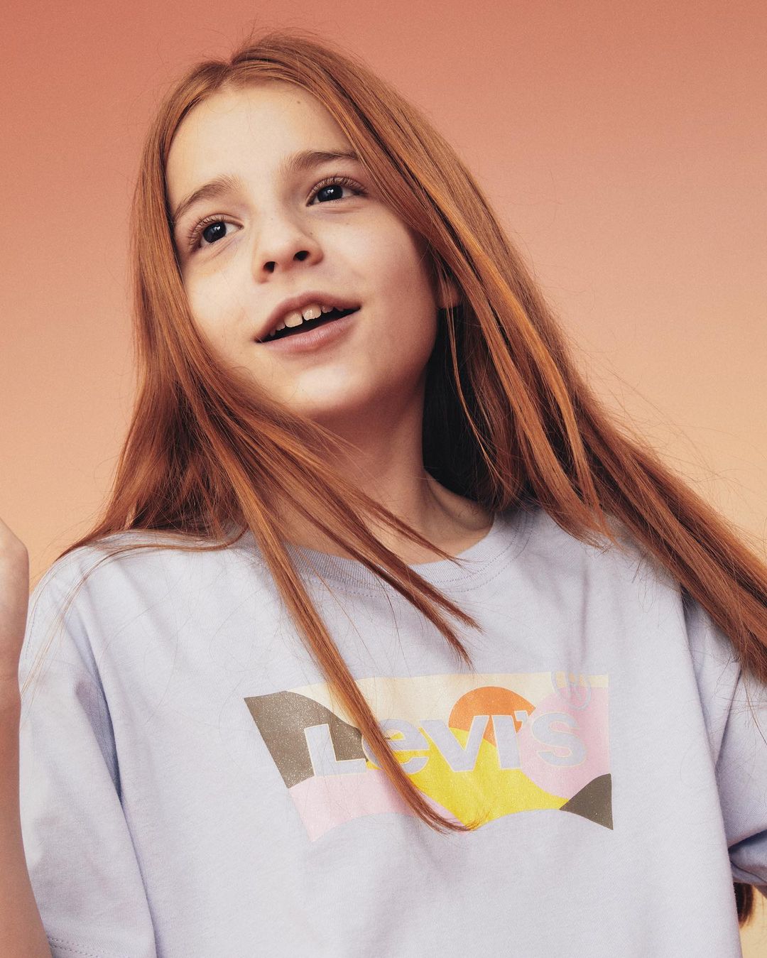 Portrait of a young female child with long red hair, wearing a light blue t-shirt with a colorful Levi's logo, looking up.