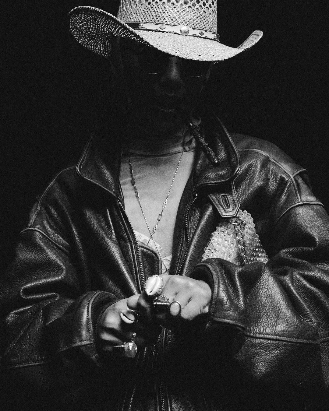 Monochrome portrait of a person in a cowboy hat and leather jacket, wearing rings and necklaces, set against a dark background.