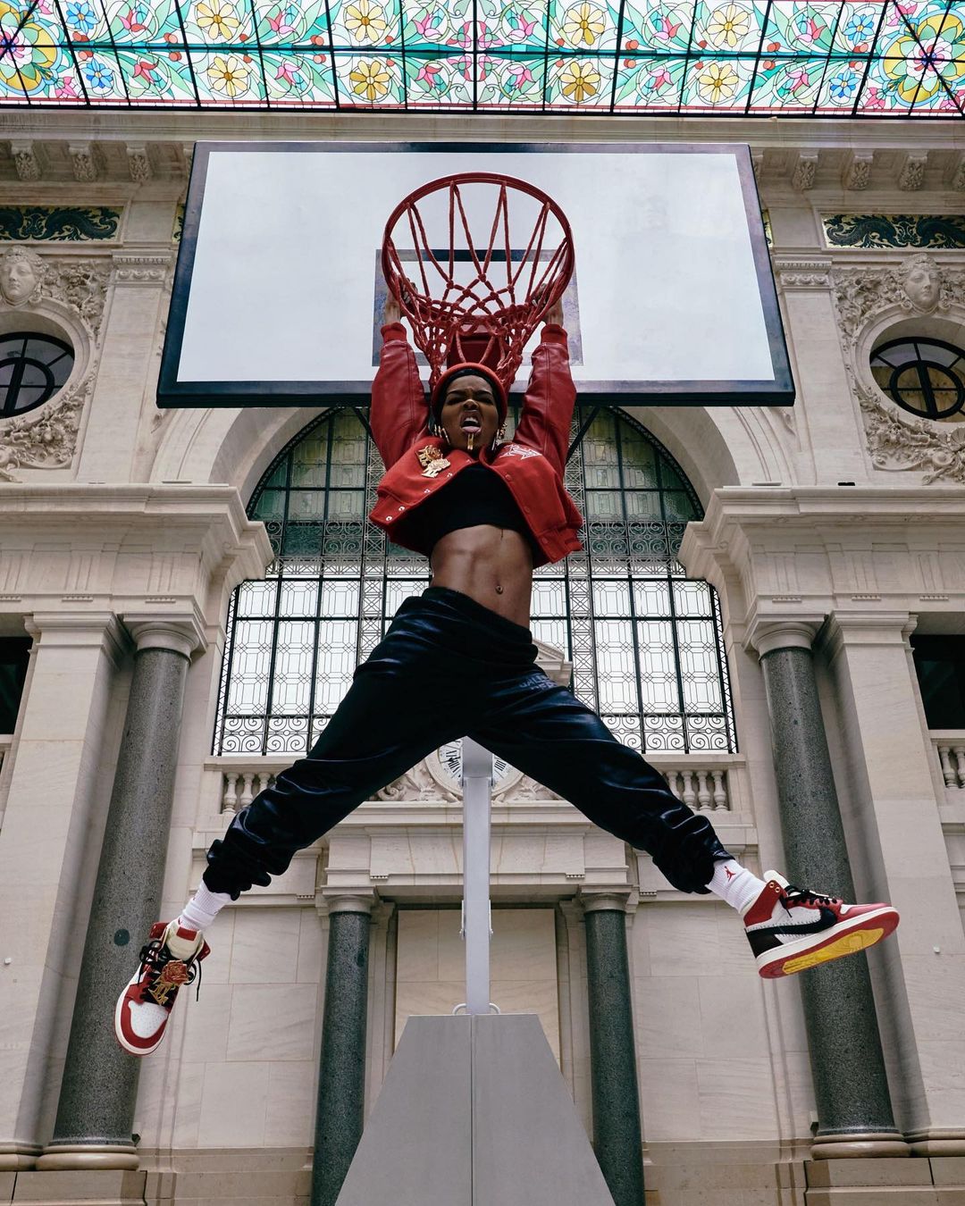 Athletic woman leaps for a basketball hoop inside a grand hall, clad in a red jacket and black pants, showcasing dynamic athleticism.